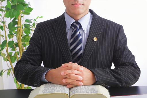 黒いマットなツヤ感のあるテーブルの前に男性が座っている  
かっちりしたスーツ姿で胸元のピンバッジが光っている  
両手を大きな法律書のような本にのせている。